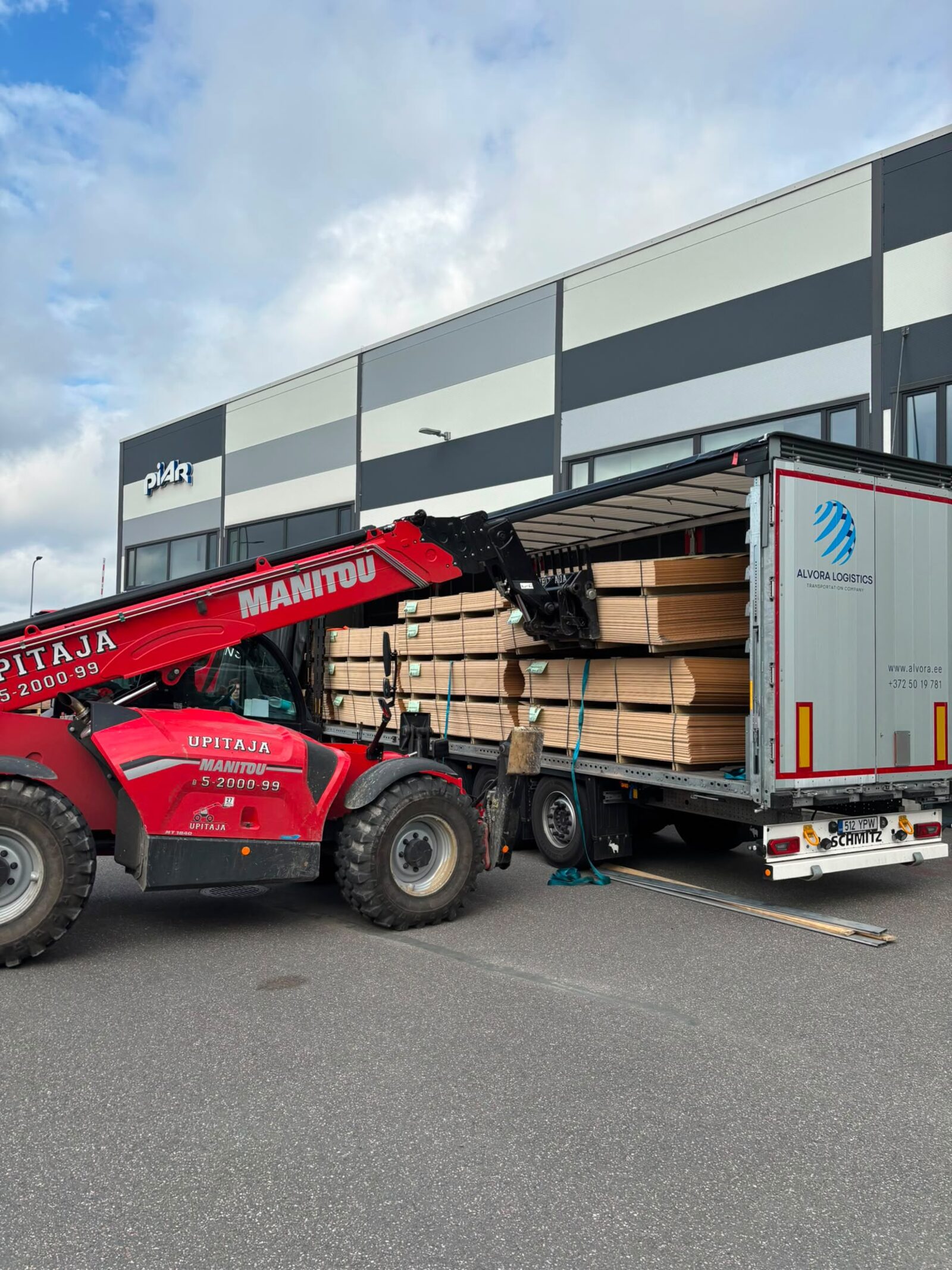Red Manitou telehandler loading an Alvora Logistics curtain-side trailer with sheet materials at a customer site